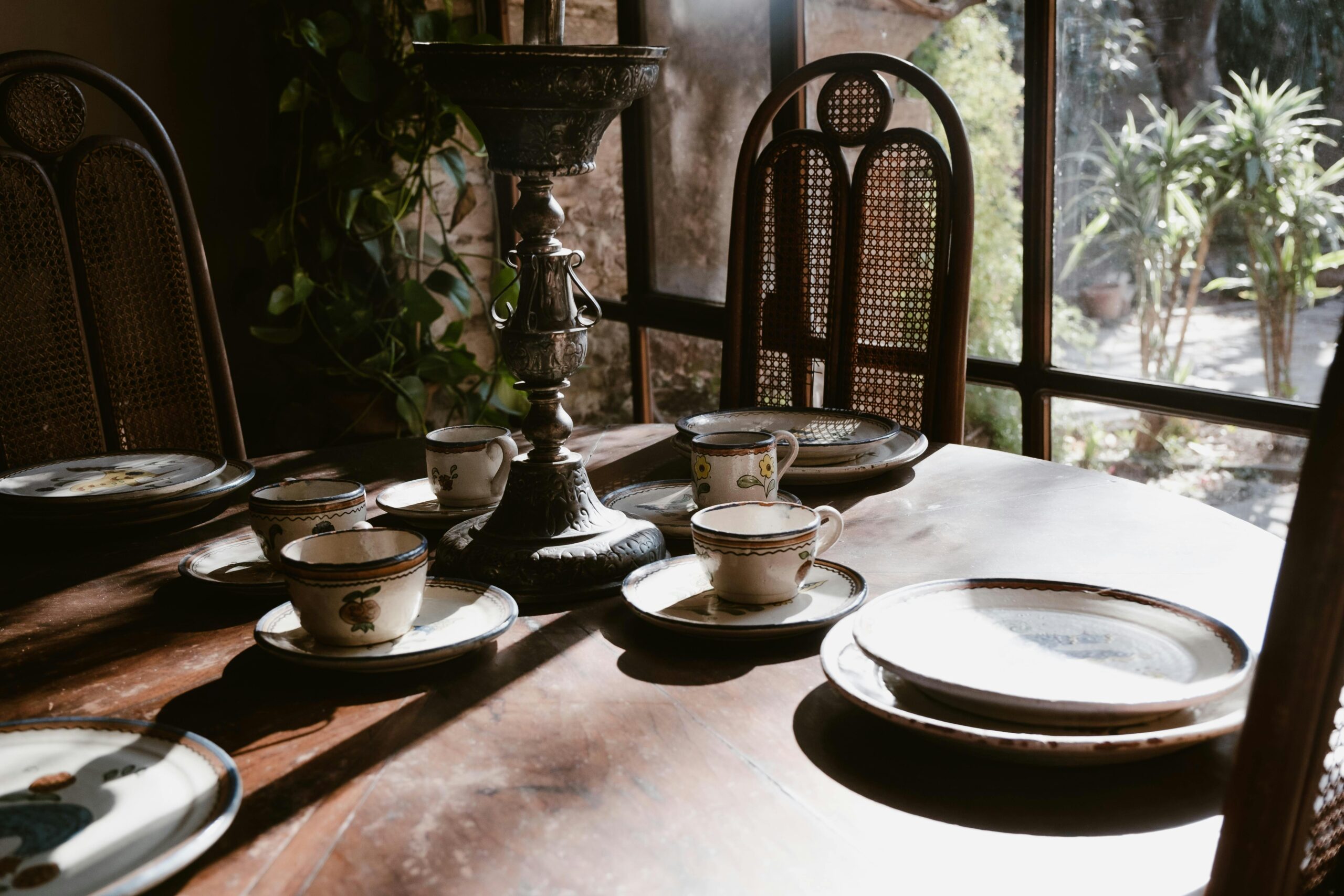 Vintage tea cups and plates set on a wooden table by a sunlit window overlooking greenery.